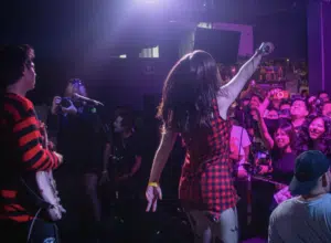 A band performs on stage with a lead singer in a red plaid dress, facing a crowd of enthusiastic fans capturing the moment with phones.